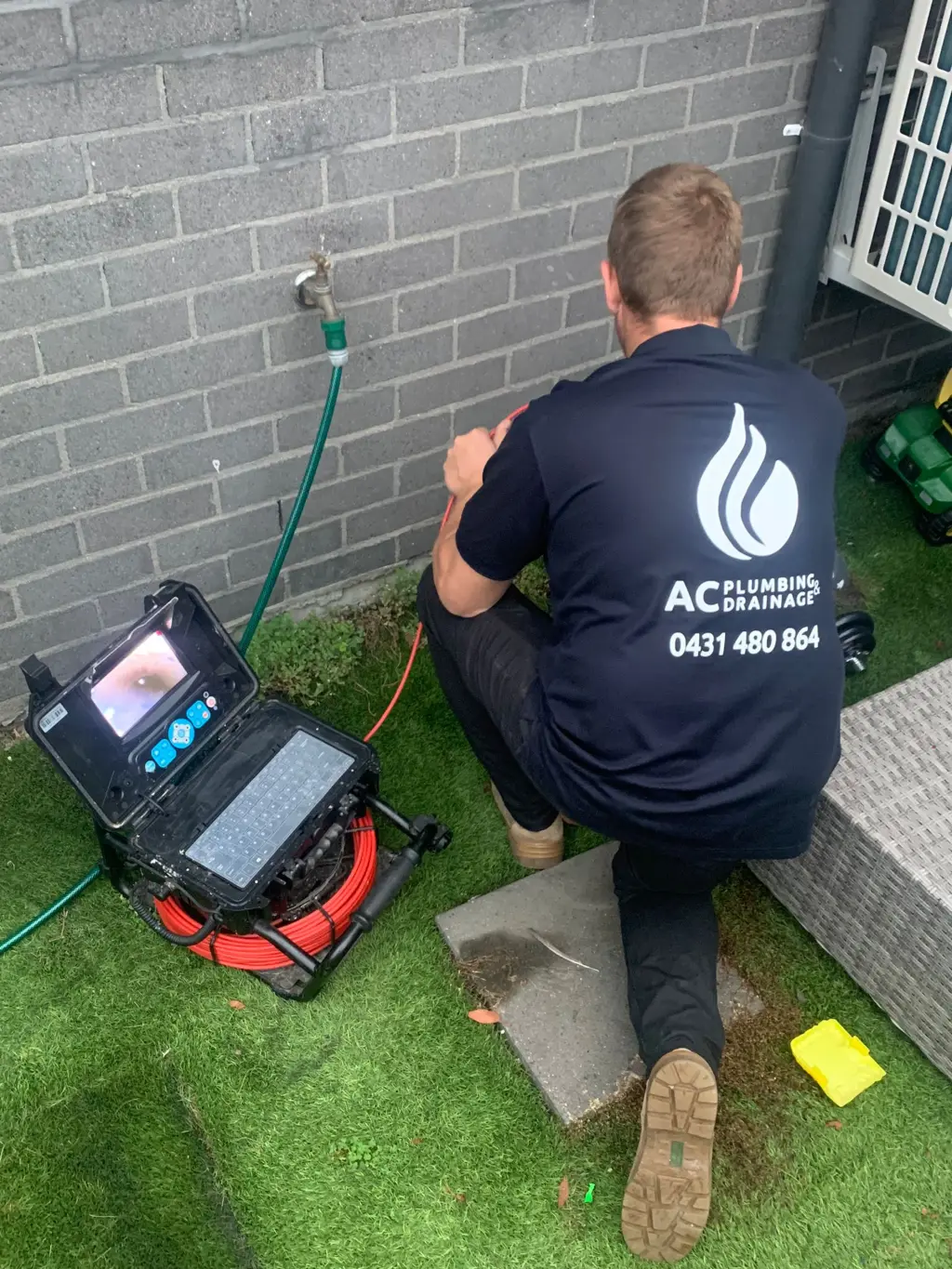 An AC Plumbing plumber running a CCTV camera reel into a drain beside a brick wall.